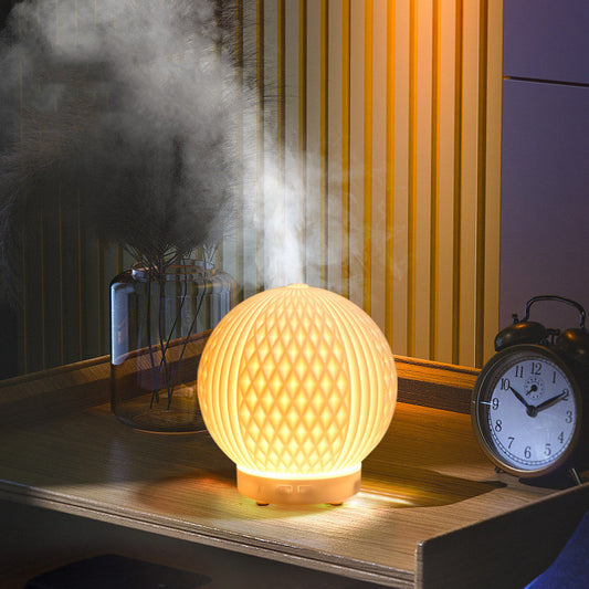 Glowing automatic essential oil diffuser emitting mist on wooden table near clock and vase