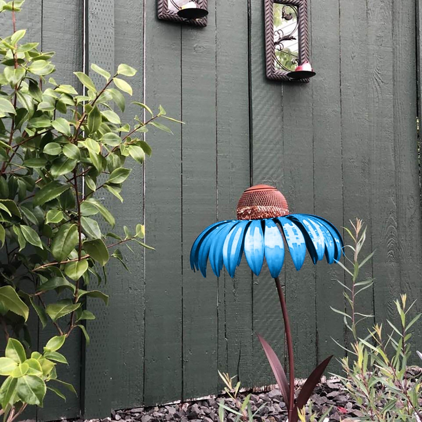 Blue metal petal shaped bird feeder with copper top in garden against green wooden fence