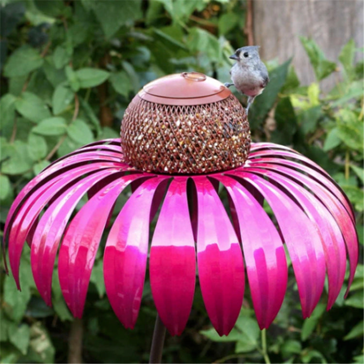 Pink metal petal shaped bird feeder with small bird perched on edge in garden setting