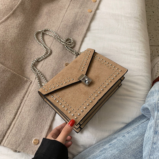 Brown studded purse with silver chain placed on beige coat and white bedsheet with jeans next to it