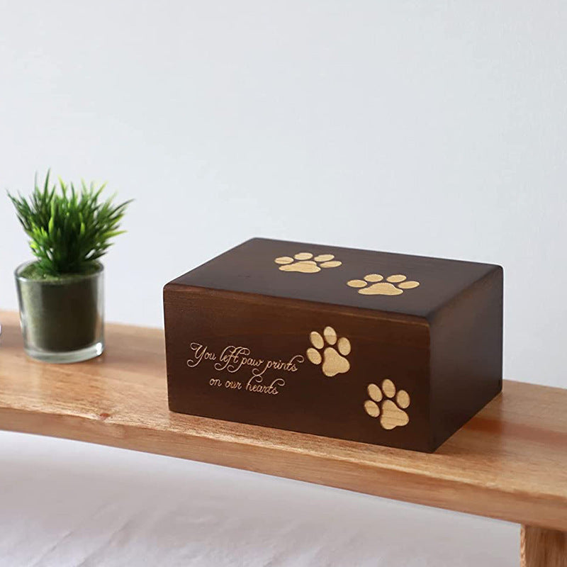 Wooden pet urn for ashes with engraved paw prints and inscription on a wooden shelf next to a small plant