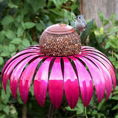 Pink metal petal shaped bird feeder with small bird perched on edge in garden setting